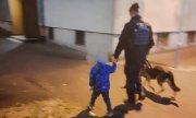 Policjant z psem służbowym idzie i trzyma za rękę dziecko - zdjęcie jest zrobione na z tyłu, niewyraźne, bo osoby się poruszają