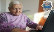 starsza pani w jasnofioletowym sweterku siedzi przed ekranem komputera. W prawym górnym rogu policyjna odznaka z białym napisem na niebieskim tle: Dolny Śląsk