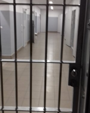 Zdjęcie kolorowe przedstawia korytarz pomieszczeń dla osób zatrzymanych.  Na przednim planie widoczne są kraty , po lewej i prawej stronie w korytarzu za kratami widać drzwi do pomieszczeń