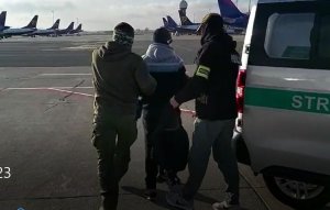 funkcjonariusze na płycie lotniska prowadzą zatrzymanego mężczyznę. W dali widoczne są samoloty