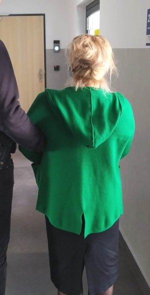 Kobieta stojąca tyłem włosy blond spięte spinką, bluza z kapturem koloru zielonego oraz czarna spódnica z rozcięciem z tyłu.