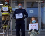 umundurowany policjant, strażak oraz dziewczynka stoją przed jednostką Policji trzymając w ręku kartki z napisami apelującymi o stosowanie się do zasad w związku z pandemią koronawirusa