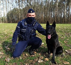 Zdjęcie przedstawia policjanta z psem służbowym. Czarny owczarek niemiecki siedzi obok swojego przewodnika