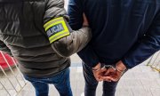 Policjant kryminalny w ubraniach cywilnych trzyma zatrzymaną osobę, która ma założone kajdanki na ręce trzymane z tyłu