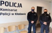 Dwóch umundurowanych policjantów stoi obok siebie w maseczkach, przy dużym logo umieszczonym na ścianie - Komisariat Policji w Kłodawie