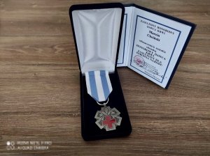Na biurku leży w pudełku medal w kształcie gwiazdy, a środku czerwony krzyż i kwiat lilii oraz biało- niebieska szarfa. Z tyłu postawiona książeczka z tekstem ZASŁUŻONY HONOROWY DAWCA KRWI Marcin Choiński odznaczony/a został/a odznaką HONOROWY DAWCA KRWI ZASŁUŻONY DLA ZDROWIA NARODU. Na dole pieczątka: Z upoważnienia MINISTRA ZDROWIA PODSEKRETARZ STANU Sławomir Gadowski oraz własnoręczny podpis. W prawym dolnym rogu: Minister Zdrowia