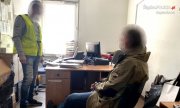 zatrzymany mężczyzna siedzi na krześle w pomieszczeniu jednostki Policji, obok stoi policjant ubrany w żółtą kamizelkę