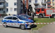radiowóz policyjny, samochody osobowe i wóz strażacki stojące przed budynkami mieszkalnymi, w głębi strażacy