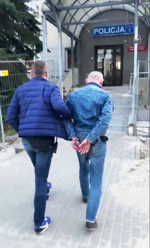 Nieumundurowany policjant prowadzi podejrzanego mężczyznę do Komisariatu Policji Warszawa Ursynów. Mężczyzna ma kajdanki założone na ręce z tyłu