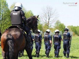 policjanci na koniach, w tle policjanci z oddziałów prewencji z tarczami w ręku stoją na trawie&quot;&gt;