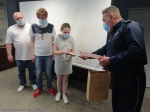 umundurowany policjant wręcza dziewczynce dyplom i prezent, obok dziewczynki stoi dwóch mężczyzn, na twarzach wszyscy mają maseczki