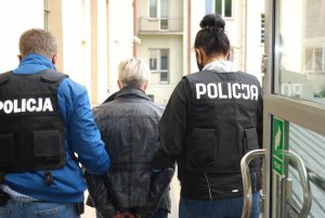 Na zdjęciu dwoje policjantów, w czarnych kamizelkach z białym napisem POLICJA, prowadzi zatrzymaną osobę