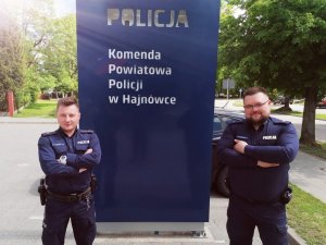 Zdjęcie przedstawia dzielnicowych. Policjanci stoją przy niebieskiej tablicy w napisem Policja, Komenda Powiatowa Policji w Hajnówce