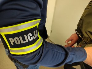 Opaska z napisem Policja na rękawie policjanta po prawej ręka zatrzymanego w kajdankach