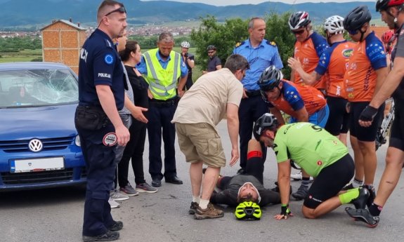 Na asfalcie leży poszkodowany cyklista. Nad nim stoją inni rowerzyści. Z lewej polski policjant, a za nim samochód uczestniczcy w wypadku.