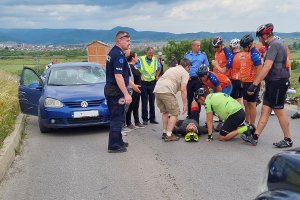 Na asfalcie leży poszkodowany cyklista. Nad nim stoją inni rowerzyści. Z lewej polski policjant, a za nim samochód uczestniczcy w wypadku.
