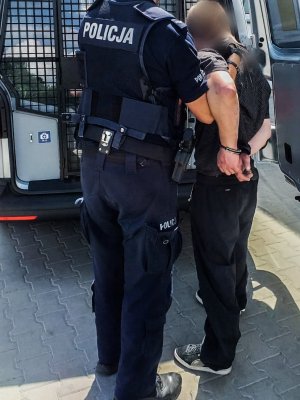 policjant trzyma zatrzymanego mężczyznę, w tle tył radiowozu
