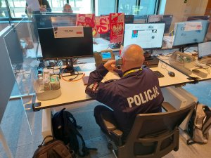 policjant przy biurku rozmawia przez telefon, w tle widać monitory komputerów, szalik z barwami narodowymi