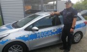 Umundurowany policjant w czapce stoi na tle policyjnego radiowozu