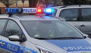 Zdjęcie kolorowe przedstawia policyjny radiowóz oznakowany z włączonymi światłami w kolorze czerwono –niebieskimi które sa umieszczone na belce