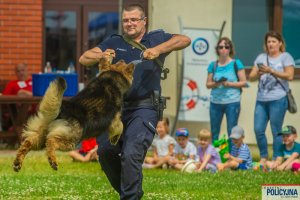 Policjant – przewodnik podczas zabawy z psem trzymającym w pysku gryzak w tle siedzące dzieci