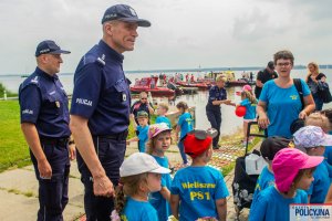 Komendant Główny Policji wraz z umundurowanymi Policjantami stojący obok grupy dzieci w tle zacumowane łodzie i motorówki