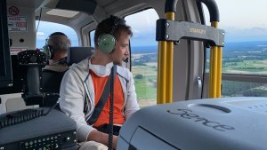 wnętrze lecącego helikoptera, na pierwszym planie siedzi mężczyzna w słuchawkach, przed nim pojemnik z napisem serce w tle pilot helikoptera