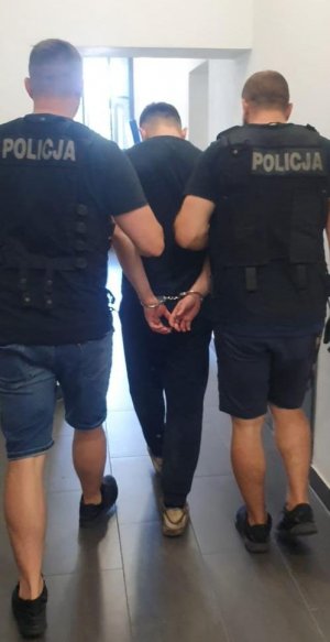 Policjanci prowadzą zatrzymanego mężczyznę, który ma założone kajdanki na ręce trzymane z tyłu