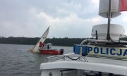 przewrócona żaglówka na jeziorze, która próbują podnieść ze swojej łodzi służby ratunkowe. Z prawej strony, z przodu widać policyjną łódź