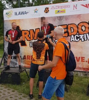 dwaj zawodnicy na podium, jeden z trzyma w dłoni puchar, drugiemu mężczyzna w pomarańczowej koszulce wręcza trofeum. Z boku widoczny jest mężczyzna, który filmuje wydarzenie trzymana w ręce kamerą.