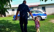 dziecko idące za rękę z policjantem, w tle budynek mieszkalny i radiowóz
