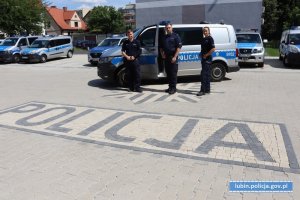 na zdjęciu trzej policjanci stoją przed radiowozem, w tle widać inne pojazdy a na ziemi napis policja