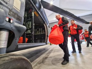 Dwóch funkcjonariuszy Państwowej Straży Pożarnej ładuje torby ze sprzętem przez drzwi desantowe śmigłowca S70i Black Hawk.