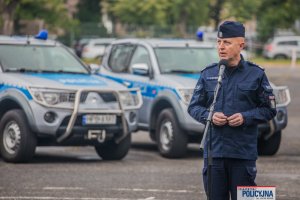 Komendant Główny Policji stoi przed mikrofonem, z tyłu widoczne radiowozy
