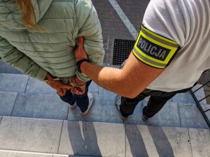 Policjant kryminalny z opaską z napisem POLICJA trzyma zatrzymaną kobietę.