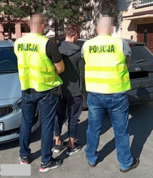 Dwóch policjantów w kamizelkach z napisem POLICJA i  zatrzymany mężczyzną, wszyscy stoją tyłem.