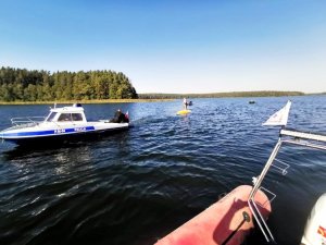 Policyjna łódź służbowa na jeziorze, w tle łódź WOPR i przewrócona żaglówka, na której jest dwóch mężczyzn&quot;&gt;