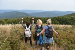 trzy kobiety z plecakami patrzą na rozpościerające się przed nimi góry, jedna z nich ma uniesione obie ręce, w których trzyma kijki, w górę. Widok z tyłu