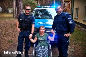 dwaj umundurowani policjanci stoją przed radiowozem z dziewczyną na wózku