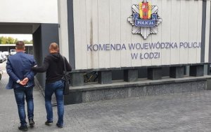 dziedziniec komendy z tablicą  z napisem Komenda Wojewódzka Policji w Łodzi, policjant po cywilnemu prowadzi zatrzymanego