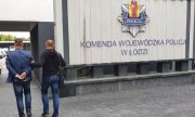 dziedziniec komendy z tablica  z napisem Komenda Wojewódzka Policji w Łodzi, policjant po cywilnemu prowadzi zatrzymanego