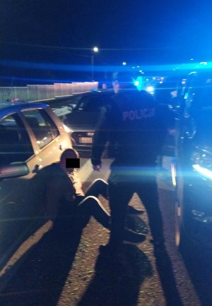Mężczyzna z kajdankami na rekach siedzi na ziemi oparty o srebrny samochód, a obok stoi policjant.