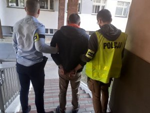Po schodach prowadzących do aresztu dwóch policjantów prowadzi zatrzymanego, który ma założone kajdanki na ręce trzymane z tyłu, jeden z nich ubrany jest w kamizelkę odblaskową z napisem policja