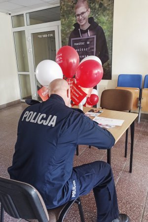 umundurowany policjant siedzi przy stoliku i wypełnia ankietę - widok z tyłu. przed nim na stoliku widać nadmuchane białe i czerwone balony oraz czerwone chorągiewki w słoiku z napisami: Zostań dawca szpiku