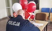 umundurowany policjant siedzi przy stoliku i wypełnia ankietę - widok z tyłu. Przed nim na stoliku widać nadmuchane białe i czerwone balony oraz czerwone chorągiewki w słoiku z napisami: Zostań dawcą szpiku
