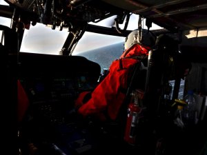 Wnętrze śmigłowca Black Hawk i widok przez jego przednią szybę na otwarte morze. Centralnie umieszczony pulpit nawigacyjny oraz siedzący przed nim funkcjonariusz w czerwonym kombinezonie i kasku.