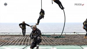 Kilku uzbrojonych funkcjonariuszy desantuje się na linach  na platformę wiertniczą na tle spokojnego morza.