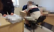 zatrzymany mężczyzna siedzi na krześle przy biurku w trakcie przesłuchania, policjanci za biurkiem notują i przekładają dokumenty