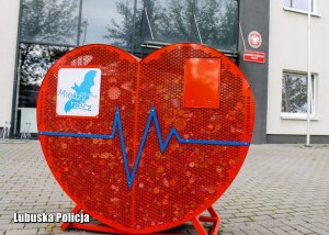 Czerwony, metalowy pojemnik w kształcie serca, a w nim plastikowe nakrętki - w tle budynek komendy Policji