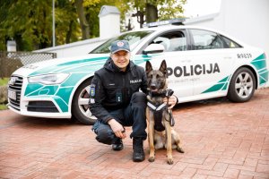 Umundurowany litewski policjant z psem służbowym kuca i pozuje do zdjęcia. Z tyłu widoczny jest policyjny radiowóz